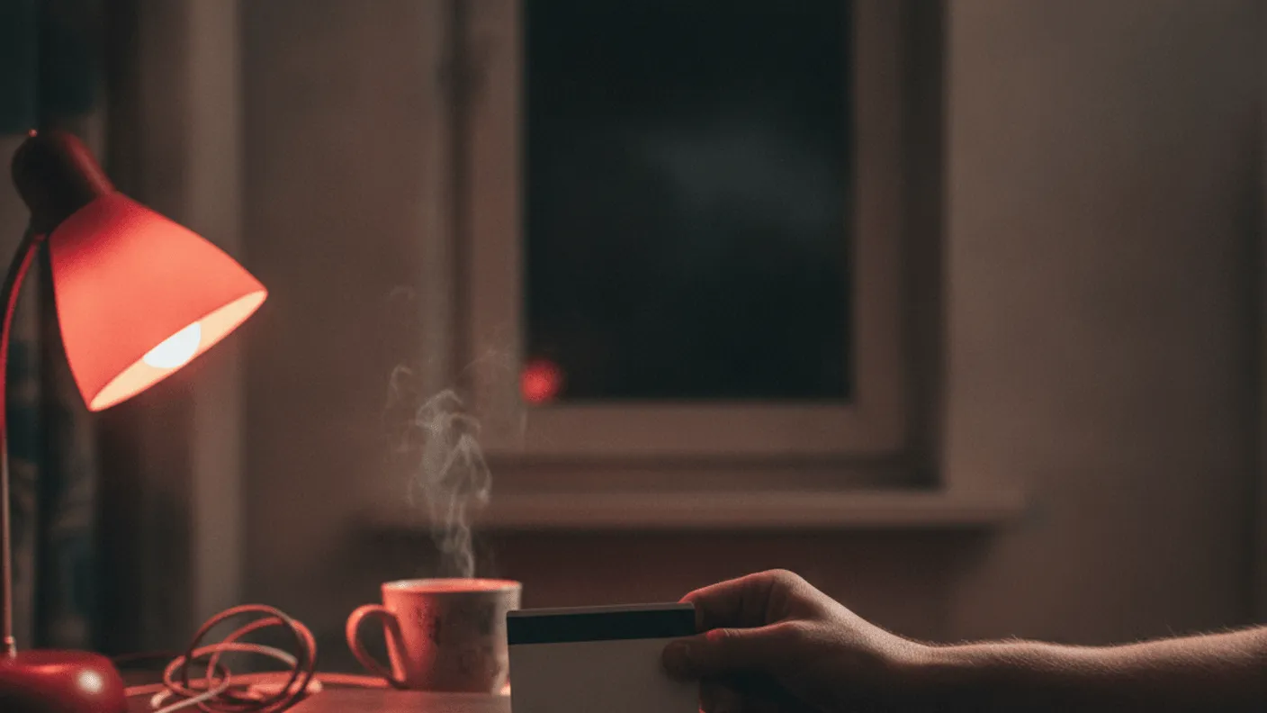 A hand holds a payment card over a laptop keyboard on a wooden desk at night with a steaming mug and red lamp.