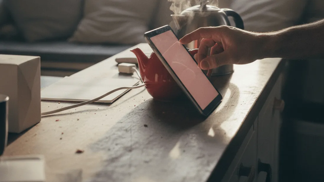 A hand adjusts a tablet propped against a red teapot, with a steaming kettle and window light in the background.