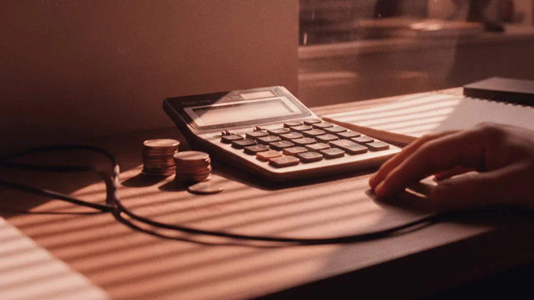 A close-up of a calendar on a wall with a circled date, a calculator, and coins on a desk with afternoon light.