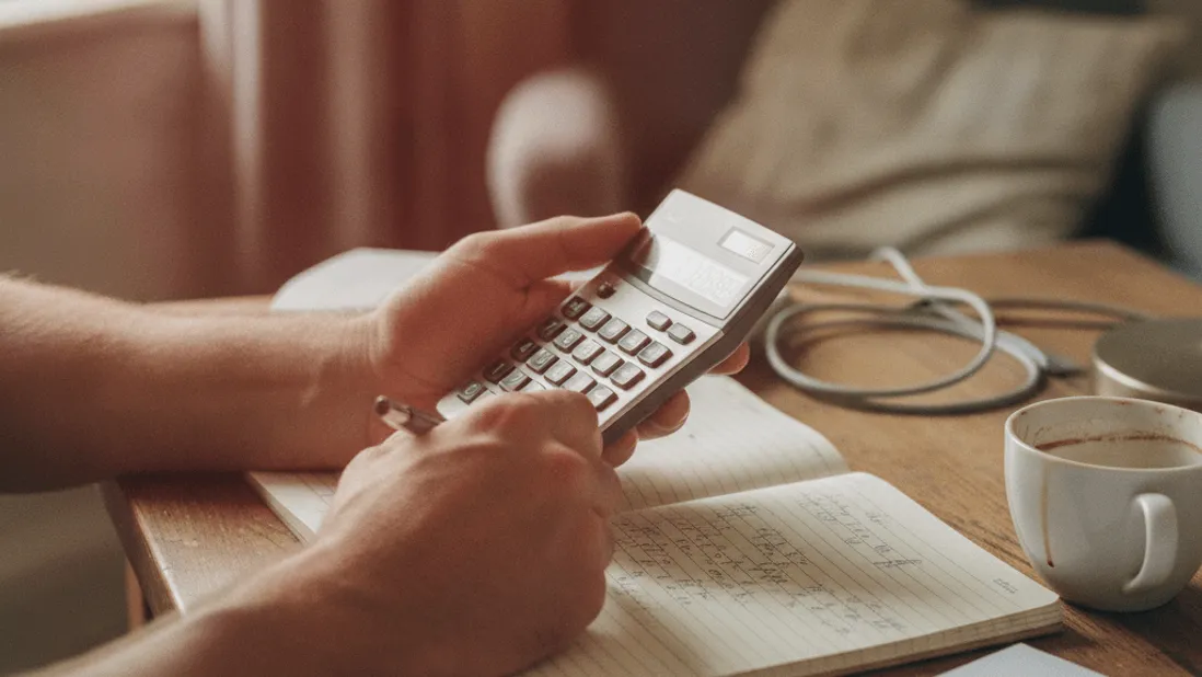 A man's forearms are shown at a desk, using a pocket calculator next to a notebook, coffee cup, and paper.