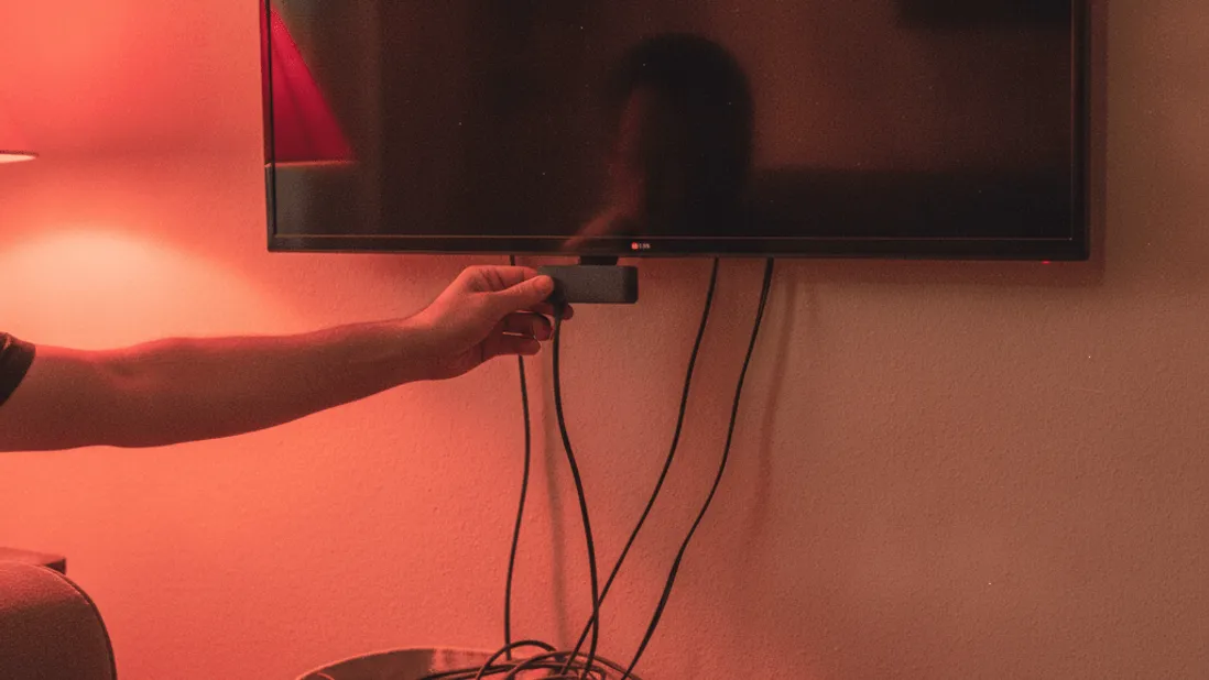 A hand plugs a streaming stick into a wall-mounted television, with cables below and red lamp light.