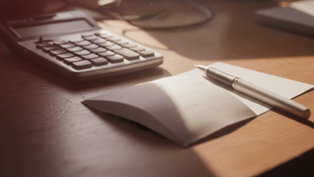 A calculator, folded receipt, and pen on a desk with morning light and a red glow.
