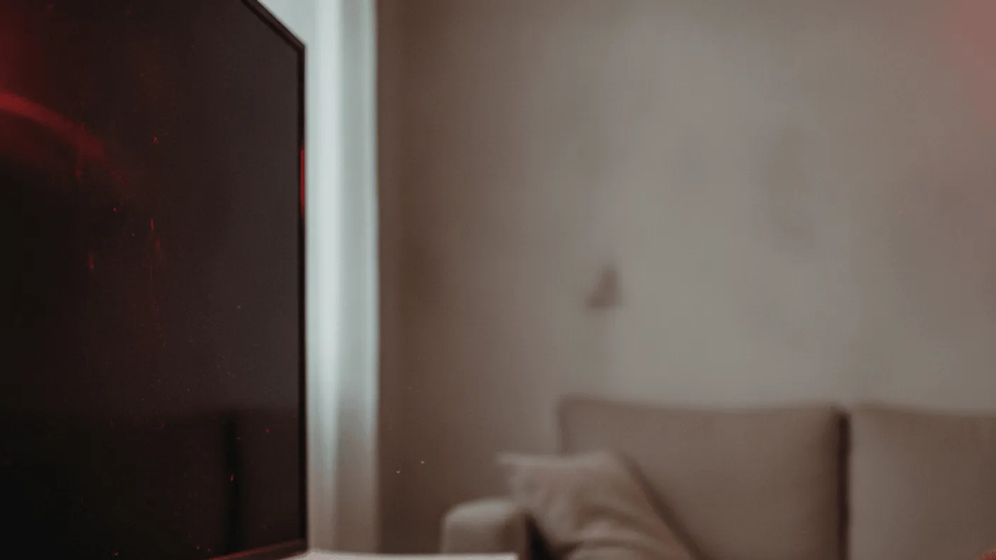 A black set-top box on a wooden shelf beside a dark television screen with a red light reflected on it.